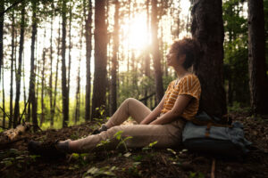 Cheerful female hiker sitting in the shade of a pine tree and resting