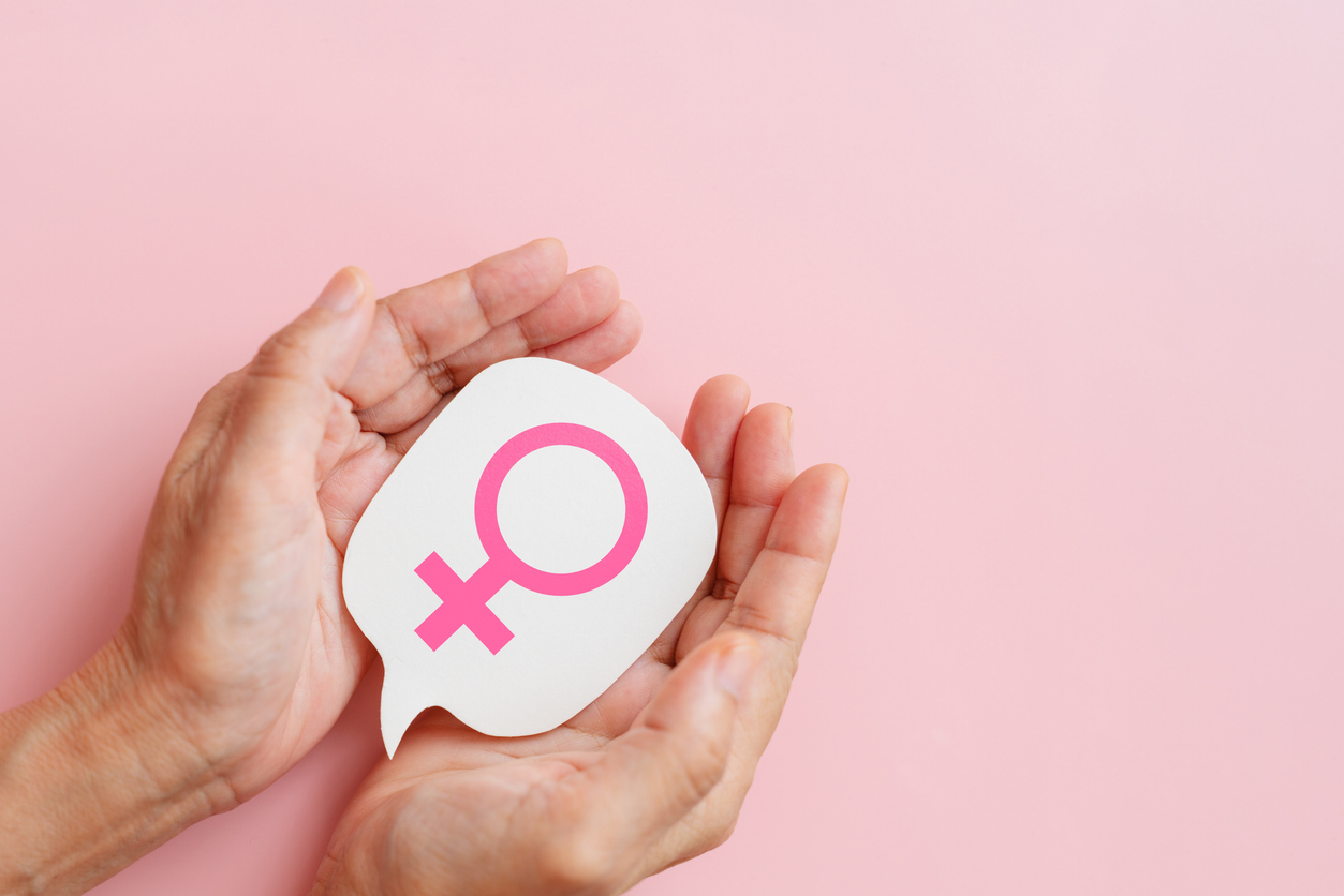 hands hold woman symbol on white paper in shape of speech bubble cut-out on pink background